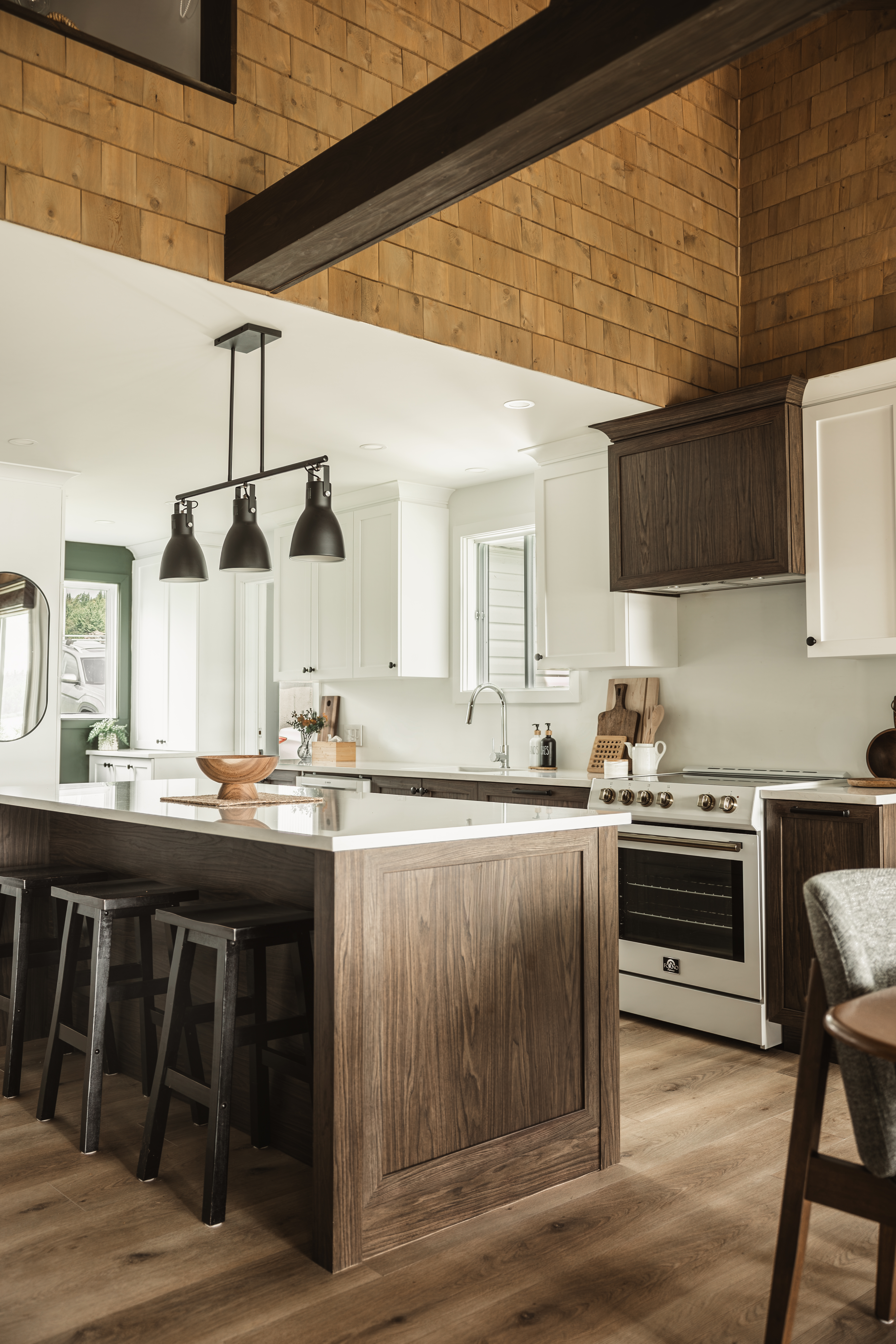 Grand îlot de cuisine en bois foncé avec tabourets et suspensions noires dans une rénovation de chalet familial. Aire ouverte conviviale conçu par la designer d'intérieur, Élévation Studio Design à Chaudière-Appalaches
