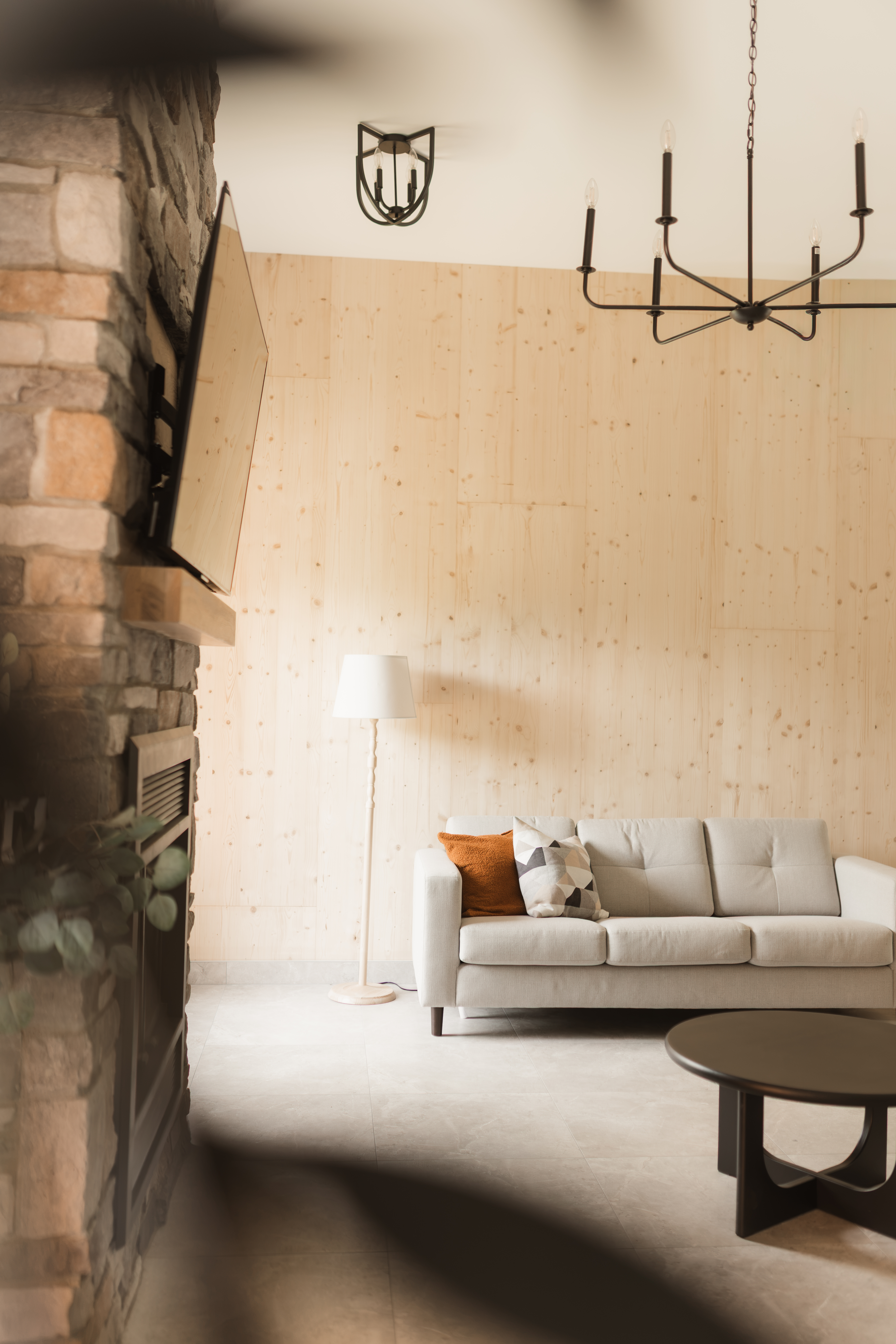 Garage transformé en salon avec foyer en pierre, mur en bois pâle, divan gris pâle et lustre noir | salon chaleureux et invitant aménagé par une designer d'intérieur