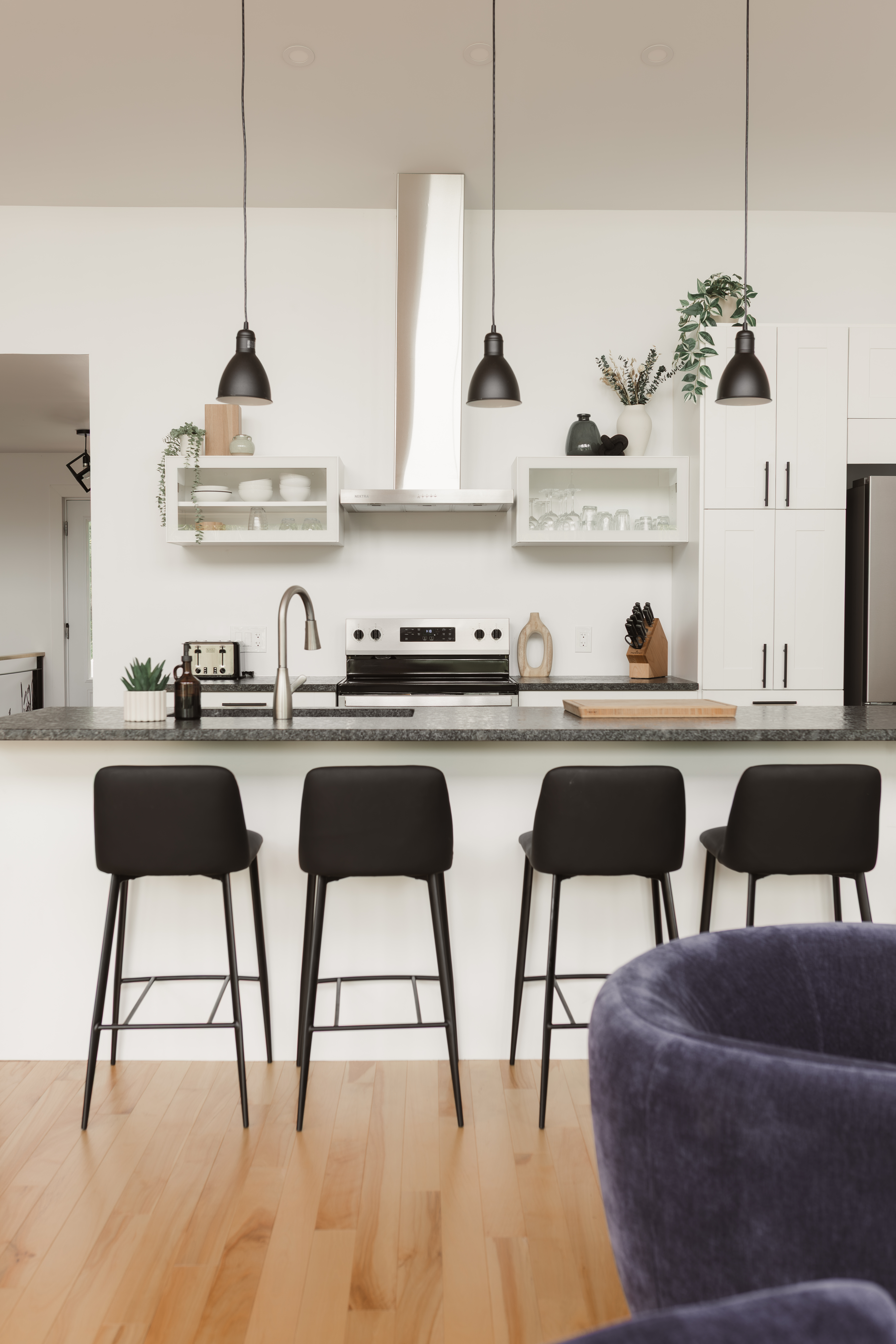 Cuisine ouverte blanche avec îlot, tabourets noirs, suspensions et plancher de bois, décorée avec soin pour accueillir les locataires | designer d'intérieur en Chaudière-Appalaches